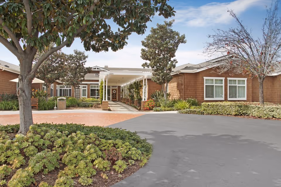 Exterior view of Ivy Park at Tustin senior living facility showing a driveway, landscaped garden with trees and shrubs, and a covered entrance to the building under a partly cloudy sky.