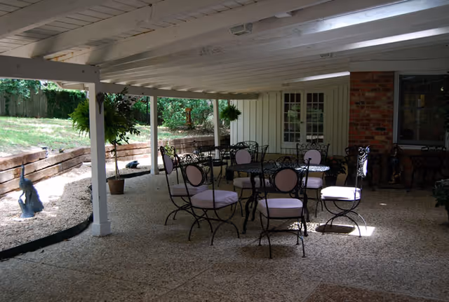 Covered outdoor patio area with several black wrought iron tables and chairs with light-colored cushions. The patio has a white wooden ceiling with beams and is adjacent to a building with white siding and a brick wall. There are potted plants and greenery visible around the patio area.