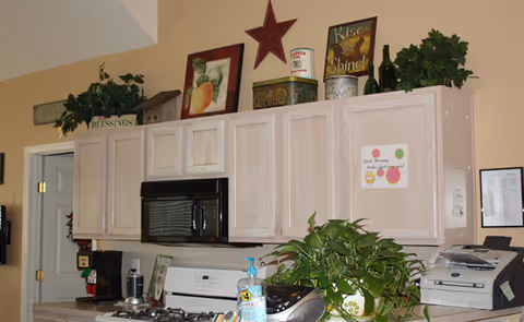 Interior view of a kitchen area with light wood cabinets, a black microwave, and a white stove. Various decorative items, including plants, framed pictures, and tins, are placed on top of the cabinets. A bottle of hand sanitizer and a green leafy plant are on the counter. A small sign with colorful circles and text is attached to one of the cabinet doors.