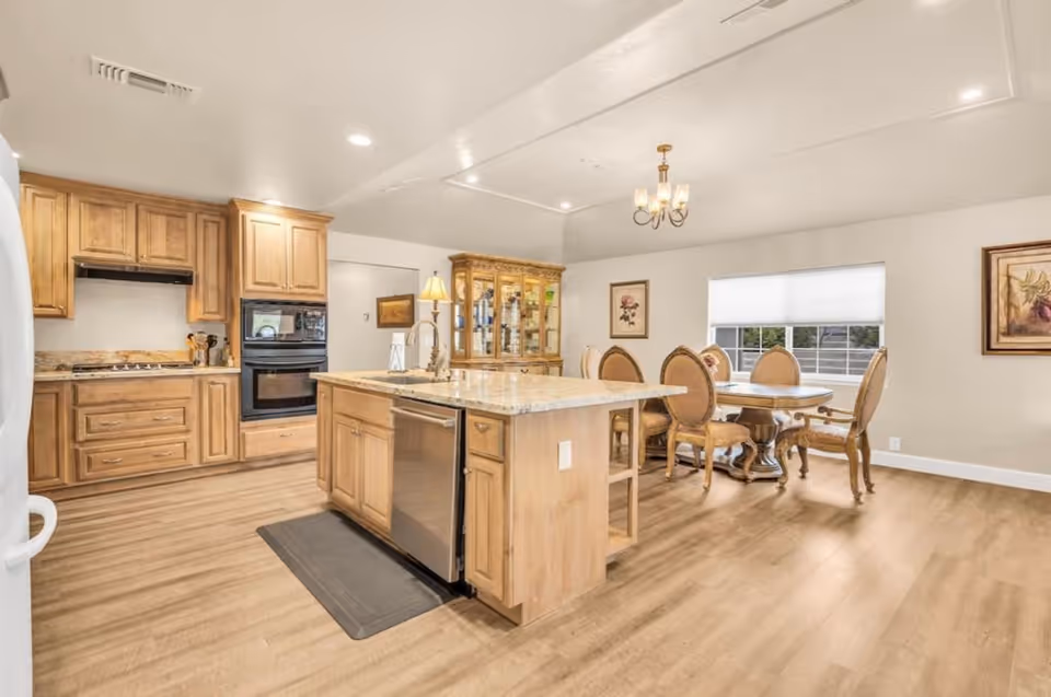 A bright and spacious kitchen and dining area featuring wooden cabinetry, a central island with a sink and dishwasher, a built-in oven, and a dining table with six upholstered chairs. The room has light-colored walls, wood flooring, a window with blinds, and decorative framed artwork on the walls.