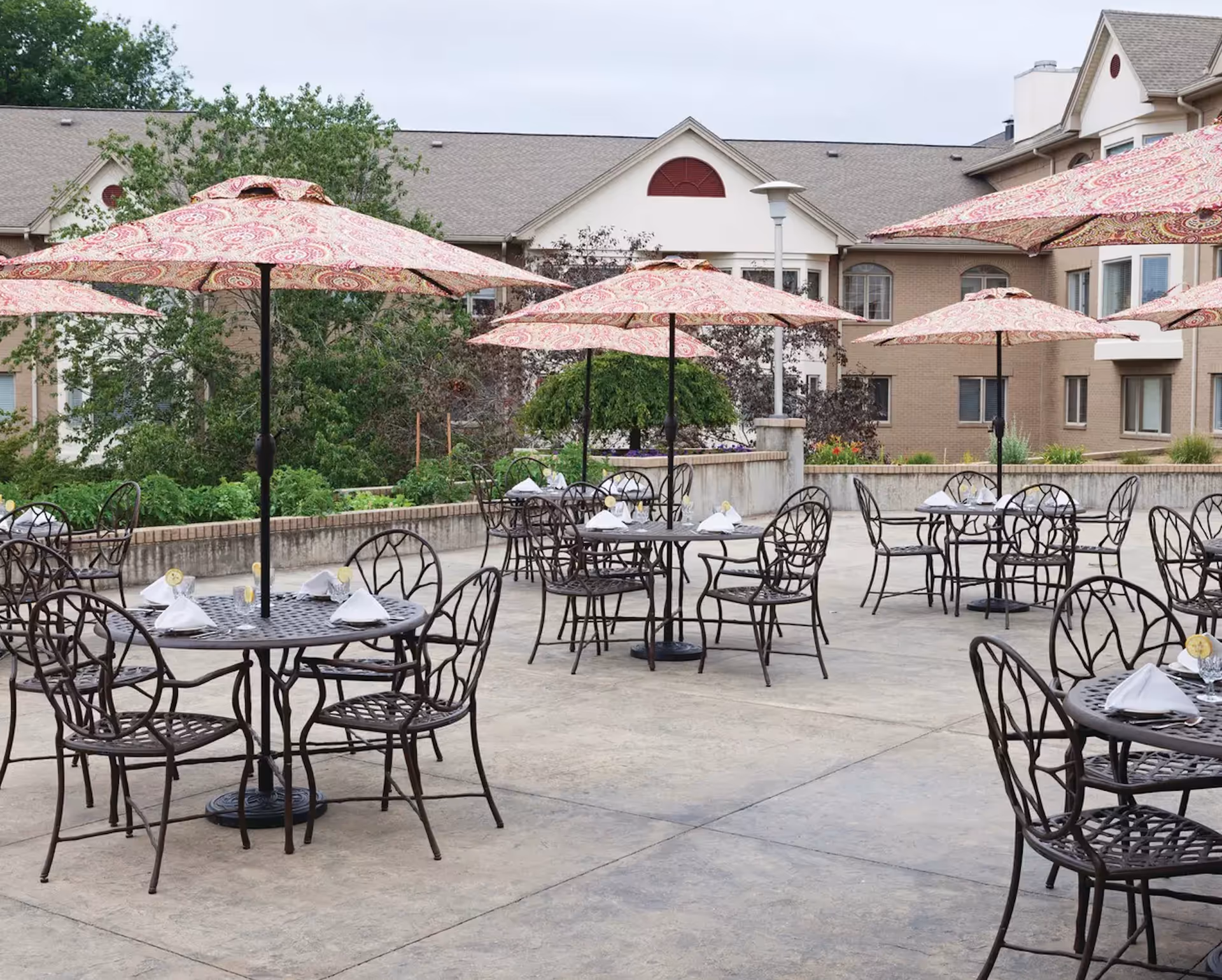 Outdoor patio area with multiple round metal tables and chairs, each table shaded by a large patterned umbrella. The tables are set with white napkins and glasses with lemon slices. In the background, there is a multi-story building with beige brick walls and windows, surrounded by greenery and trees.