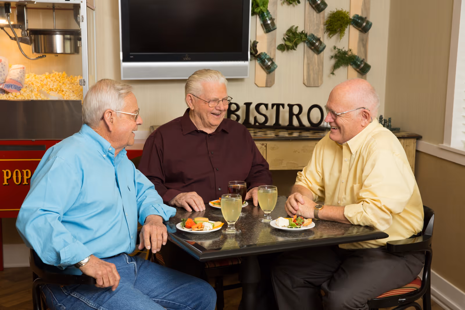 Three older men sit and laugh around a small bistro table with drinks and plates of food in a dining area.
