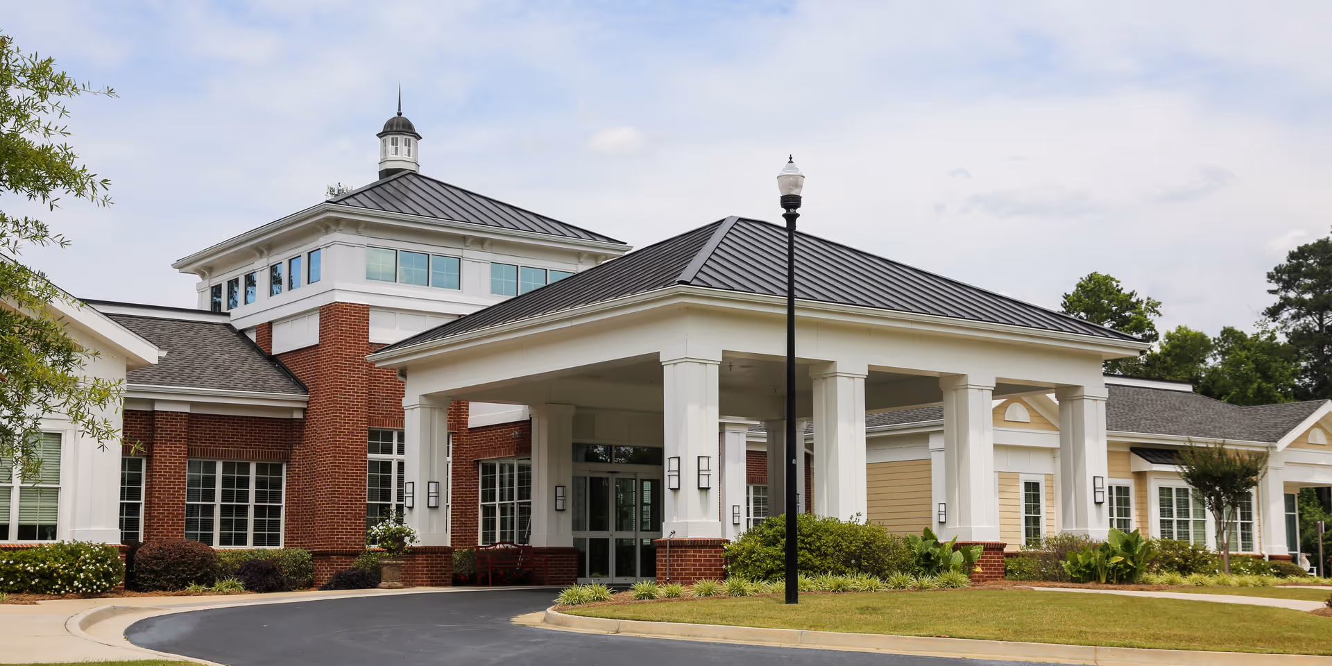 Front exterior view of Zebulon Park Health & Rehabilitation facility showing a large entrance with white columns, a covered drop-off area, brick and beige siding, multiple windows, and a landscaped lawn with bushes and a streetlamp.