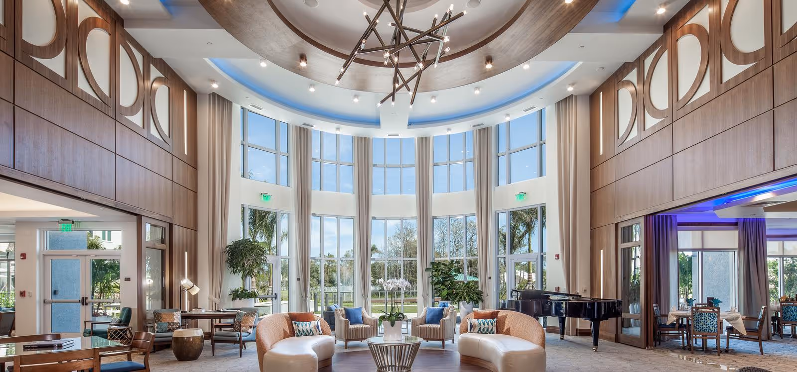 Spacious and elegant senior living facility common area with large floor-to-ceiling windows allowing natural light, modern circular ceiling design with chandelier, curved beige sofas, armchairs, potted plants, and a black grand piano near a dining area with tables and chairs.