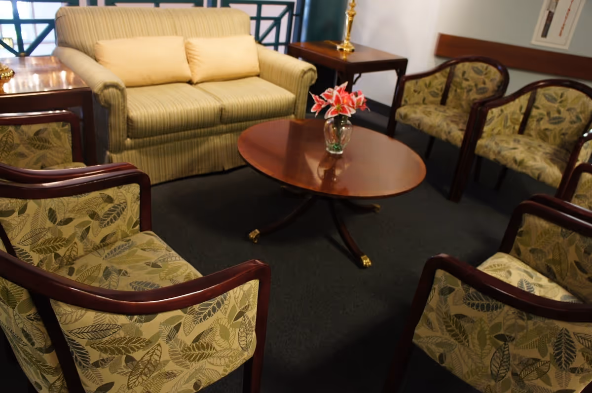 A cozy seating area with a striped loveseat with two beige cushions, surrounded by six armchairs upholstered in green leaf-patterned fabric. In the center is a wooden oval coffee table with a glass vase holding pink lilies. There are side tables with lamps in the background.