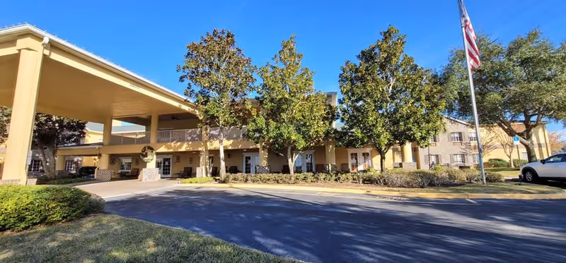 Front entrance of Hunt Trace Senior Living showing a covered porte-cochere, landscaping, and an American flag.
