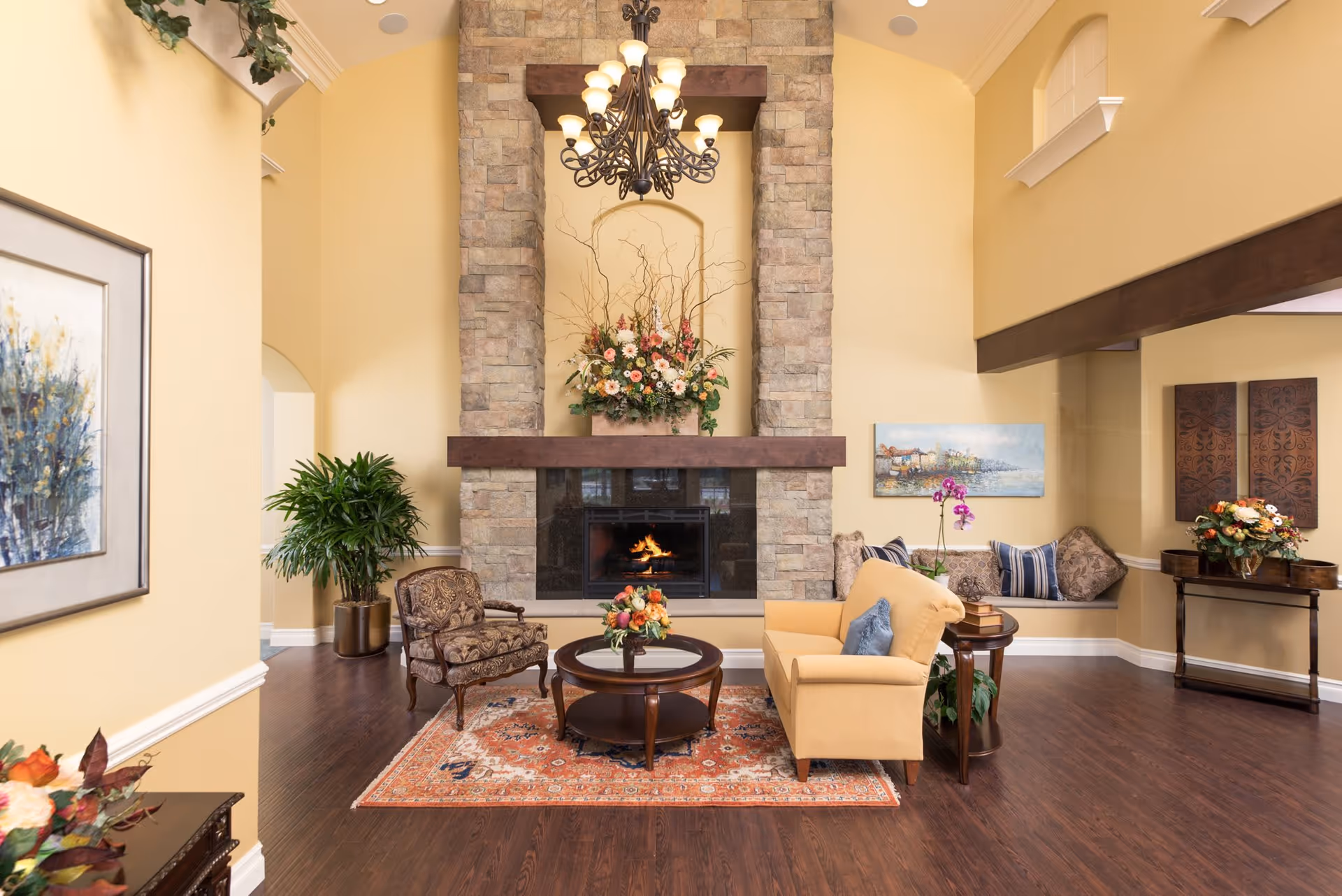 Bright communal living room with a stone fireplace, chandelier, sofa and chairs arranged around a coffee table, and decorative plants and artwork.