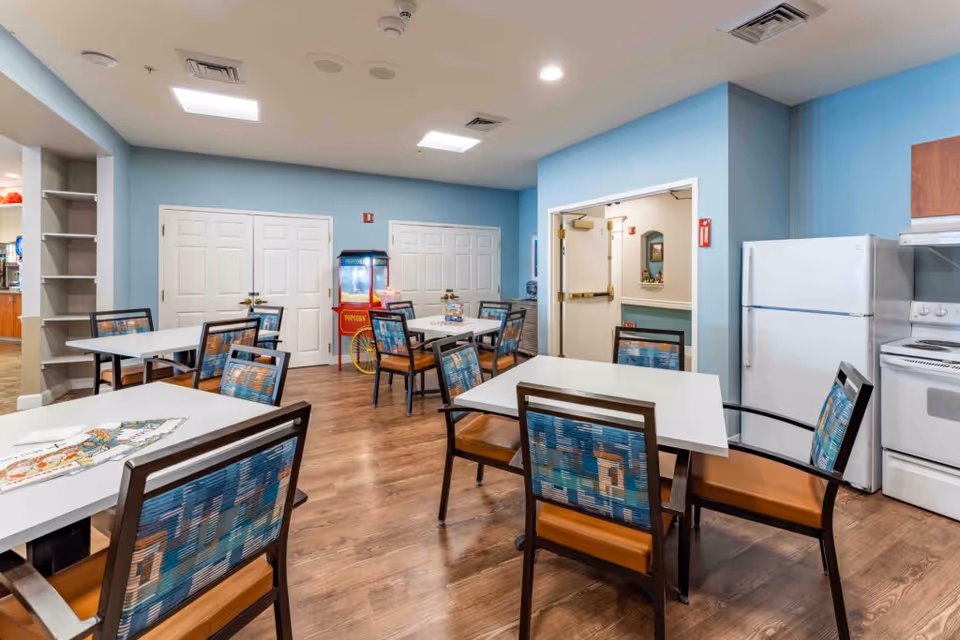 A dining area with several square white tables and chairs with patterned backs and brown seats. The room has light blue walls, wooden flooring, and ceiling lights. There is a white refrigerator and stove on the right side, and a popcorn machine against the far wall near double doors.