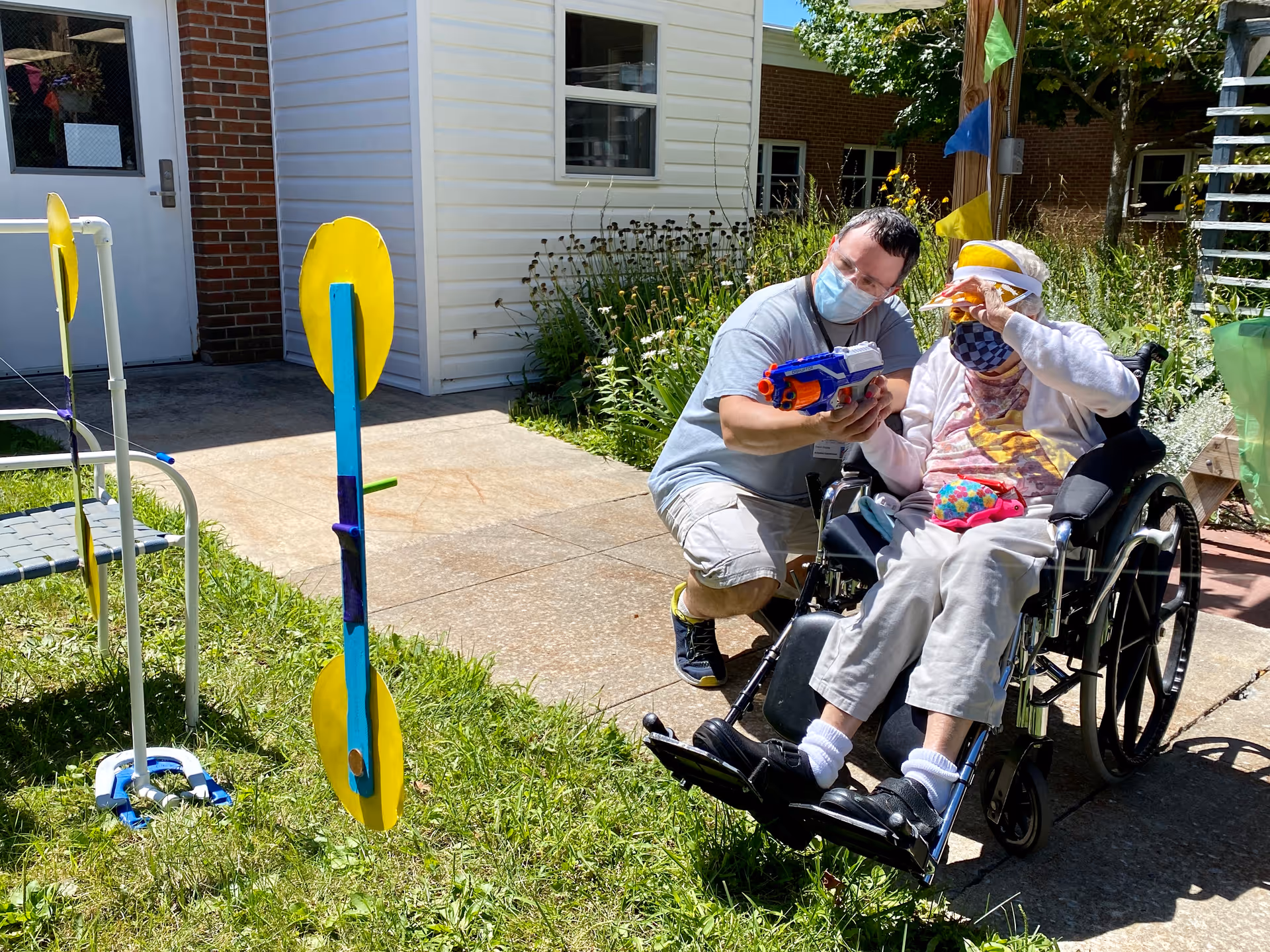 An elderly person in a wheelchair wearing a face mask and visor is outdoors on a sunny day, holding a toy gun with the assistance of a man kneeling beside them who is also wearing a face mask. They are in a garden area with grass, a concrete path, and some colorful targets set up nearby.
