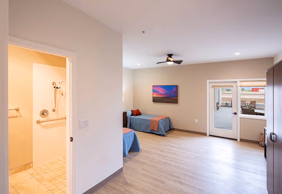 A spacious assisted living bedroom with two single beds covered in blue bedspreads and orange throws. The room has light-colored walls and wood flooring. A ceiling fan with a light is mounted on the ceiling. There is a framed picture of a sunset on the wall. A door and window lead to an outdoor patio area with seating. To the left, an open door reveals a bathroom with grab bars and a handheld showerhead.