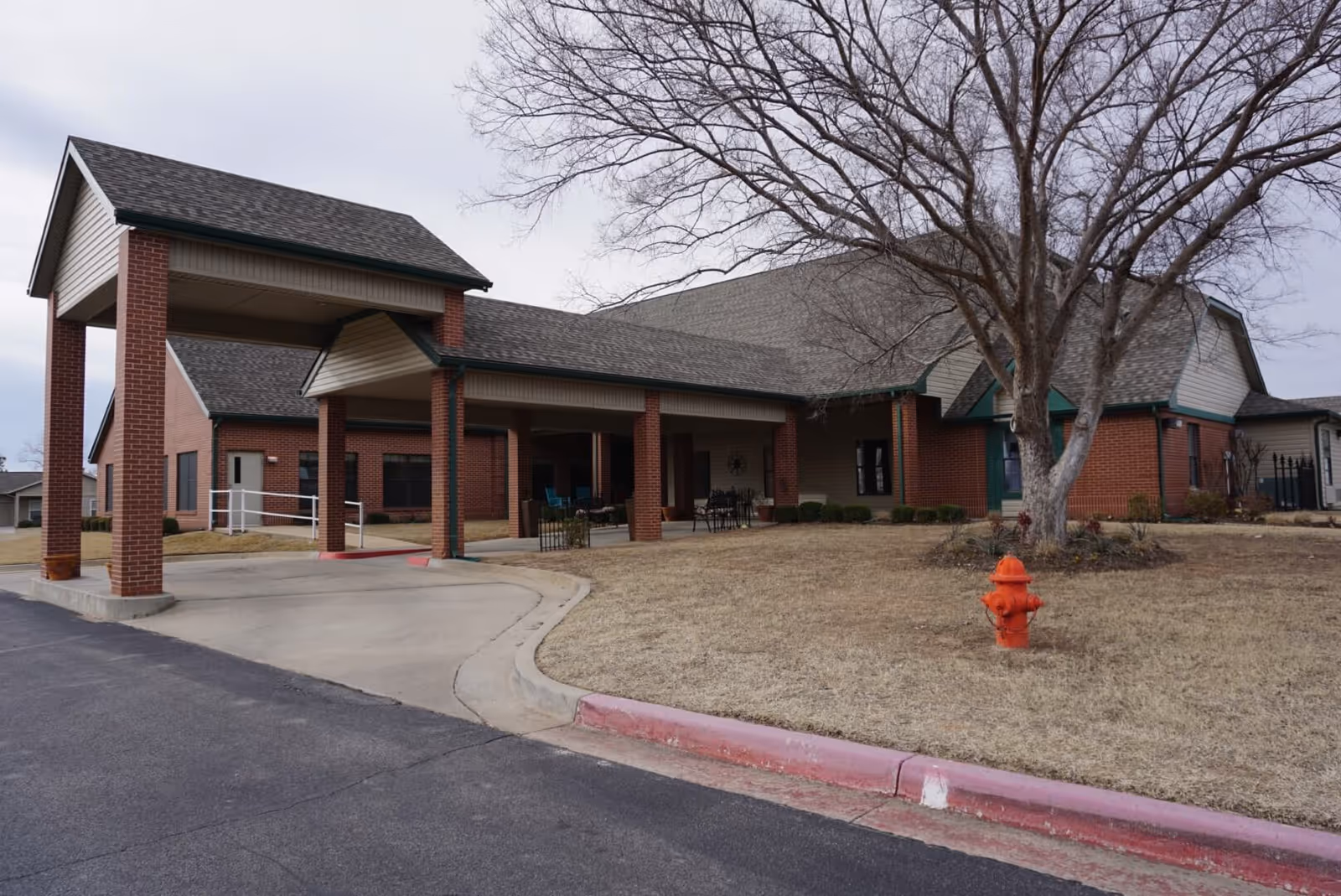 Covered entrance and drive of a single-story brick senior living building with a leafless tree and an orange fire hydrant on the front lawn.