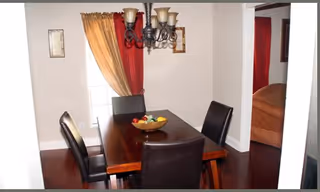 A dining room with a dark wooden rectangular table surrounded by four black leather chairs. A decorative chandelier with five lights hangs above the table. The room has light-colored walls, a window with two-tone curtains in gold and red, and framed artwork on the walls. A glimpse of an adjacent room with a brown sofa is visible through an open doorway.