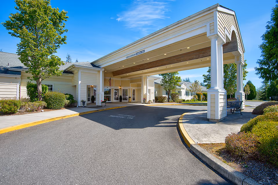 Entrance of Inland Point Retirement Community showing a covered driveway with white columns, a bench on the right side, and surrounding greenery under a clear blue sky.