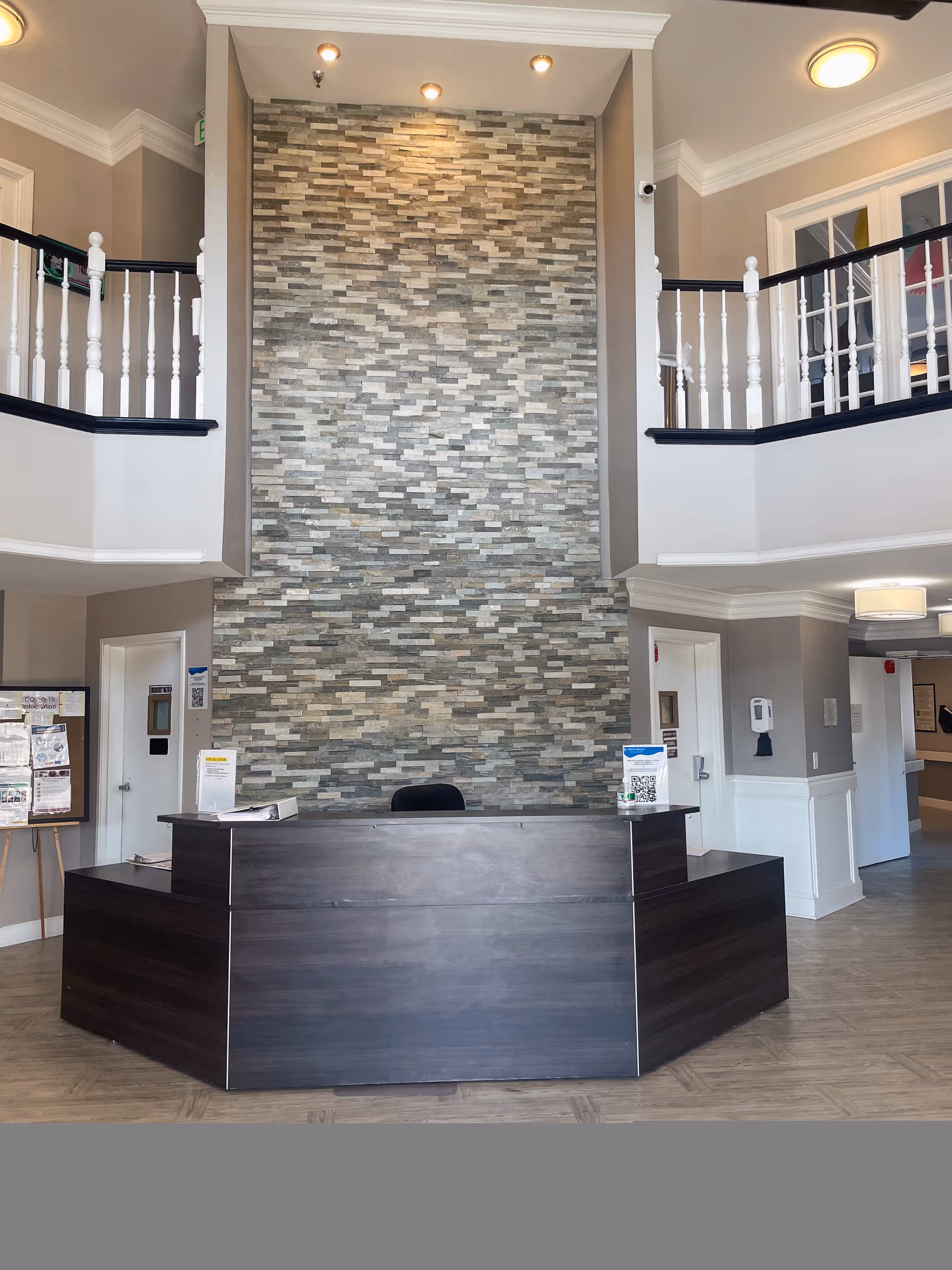Reception desk in a two-story lobby with a stacked-stone accent wall and upper balcony railings.