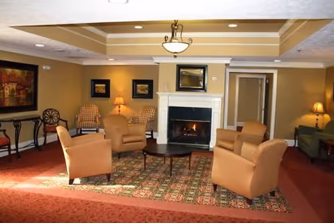 A cozy living room area with four tan armchairs arranged around a dark wooden coffee table on a patterned rug. There is a white fireplace with a fire burning and a mirror above it. The walls are painted beige and decorated with framed artwork. Two table lamps provide warm lighting, and the ceiling has recessed lighting and a central chandelier.