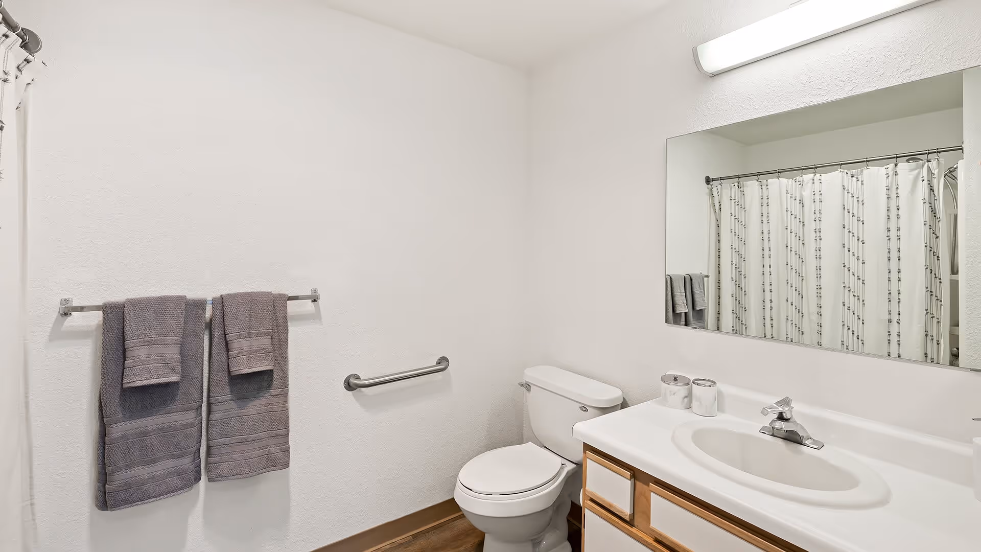 A clean bathroom with white walls featuring a toilet, a sink with a countertop and wooden cabinets underneath, a large mirror above the sink, a towel rack with two gray towels, a grab bar next to the toilet, and a shower with a white curtain patterned with vertical lines.