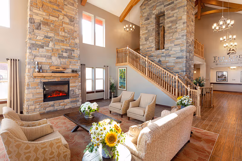 Spacious senior living lobby with a stone fireplace, seating area, and a wooden staircase under high ceilings.