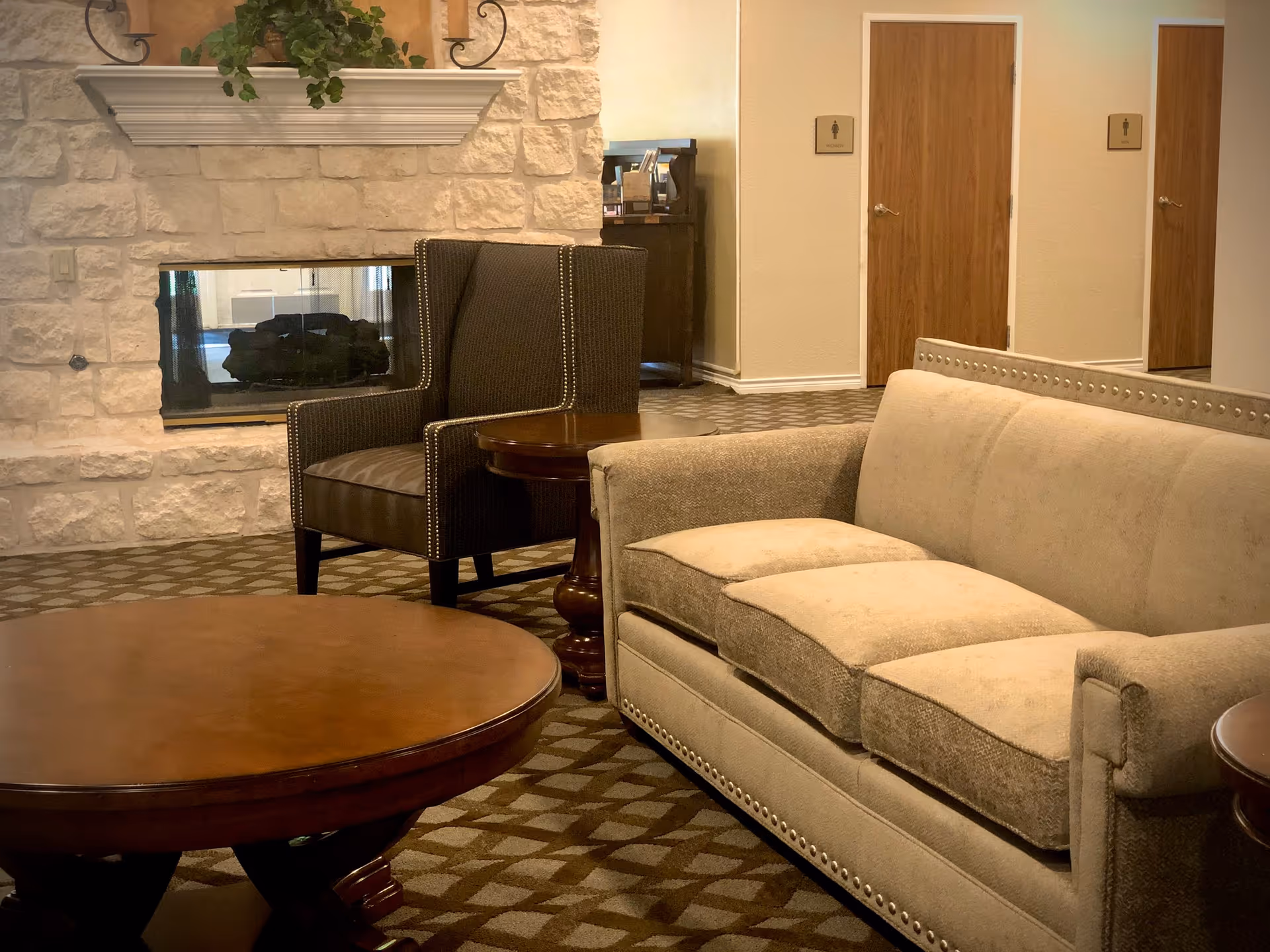 A cozy sitting area with a beige sofa and a dark upholstered armchair next to a stone fireplace. There is a round wooden coffee table in the foreground and a small wooden side table between the sofa and armchair. In the background, there are two wooden doors with signs indicating women's and men's restrooms.