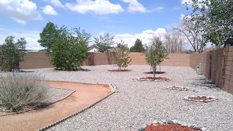 A fenced outdoor garden area with gravel ground cover, several small trees, and circular stone-bordered planting beds. The sky is partly cloudy with some blue visible.