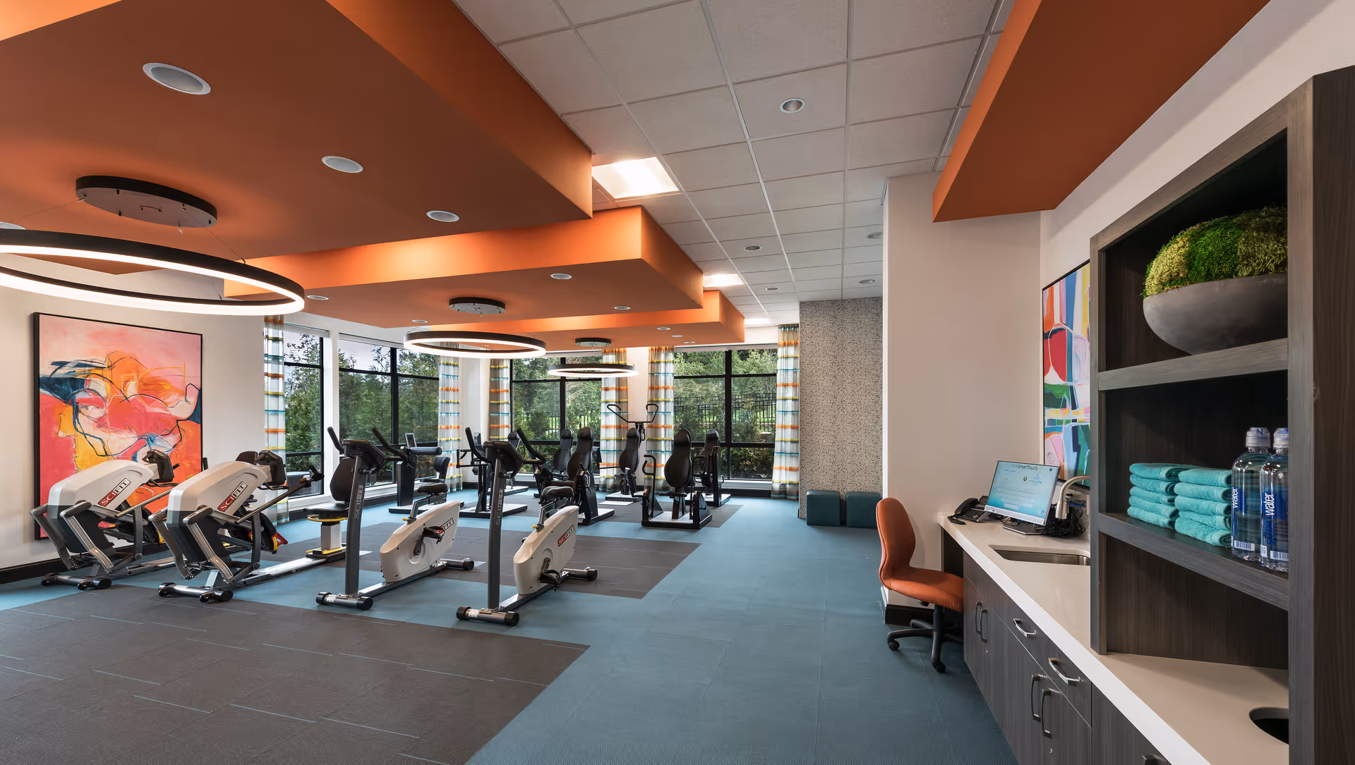 A modern fitness room with several exercise bikes and elliptical machines arranged on a blue and gray carpeted floor. The ceiling features orange beams with circular light fixtures. Large windows with striped curtains allow natural light to fill the room. On the right side, there is a desk with a computer and an orange chair, along with shelves holding neatly folded turquoise towels and water bottles. Colorful abstract artwork is displayed on the walls.