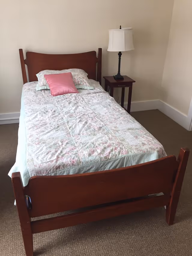 A single wooden bed with a floral quilt and a pink pillow, next to a small wooden nightstand with a white lampshade in a simple room with beige walls and carpeted floor.