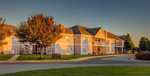 Two-story senior living building with balconies, trees, a bench, and a curved driveway bathed in warm sunset light.
