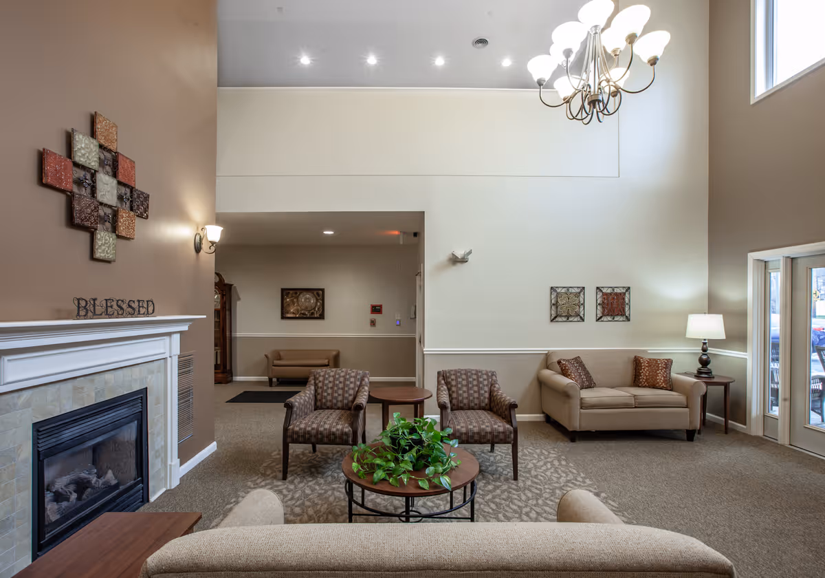 A cozy living room area with beige walls and carpet. The room features a fireplace with a decorative 'BLESSED' sign on the mantle, two patterned armchairs, a beige sofa with two pillows, a round coffee table with a green plant, and a side table with a lamp. There is a chandelier hanging from the high ceiling and wall art decorating the walls. Large windows and glass doors allow natural light into the space.