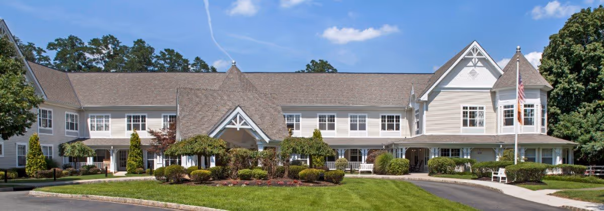 Front exterior of a two-story senior living facility with pitched roofs, a covered entrance, landscaped lawn and an American flag.