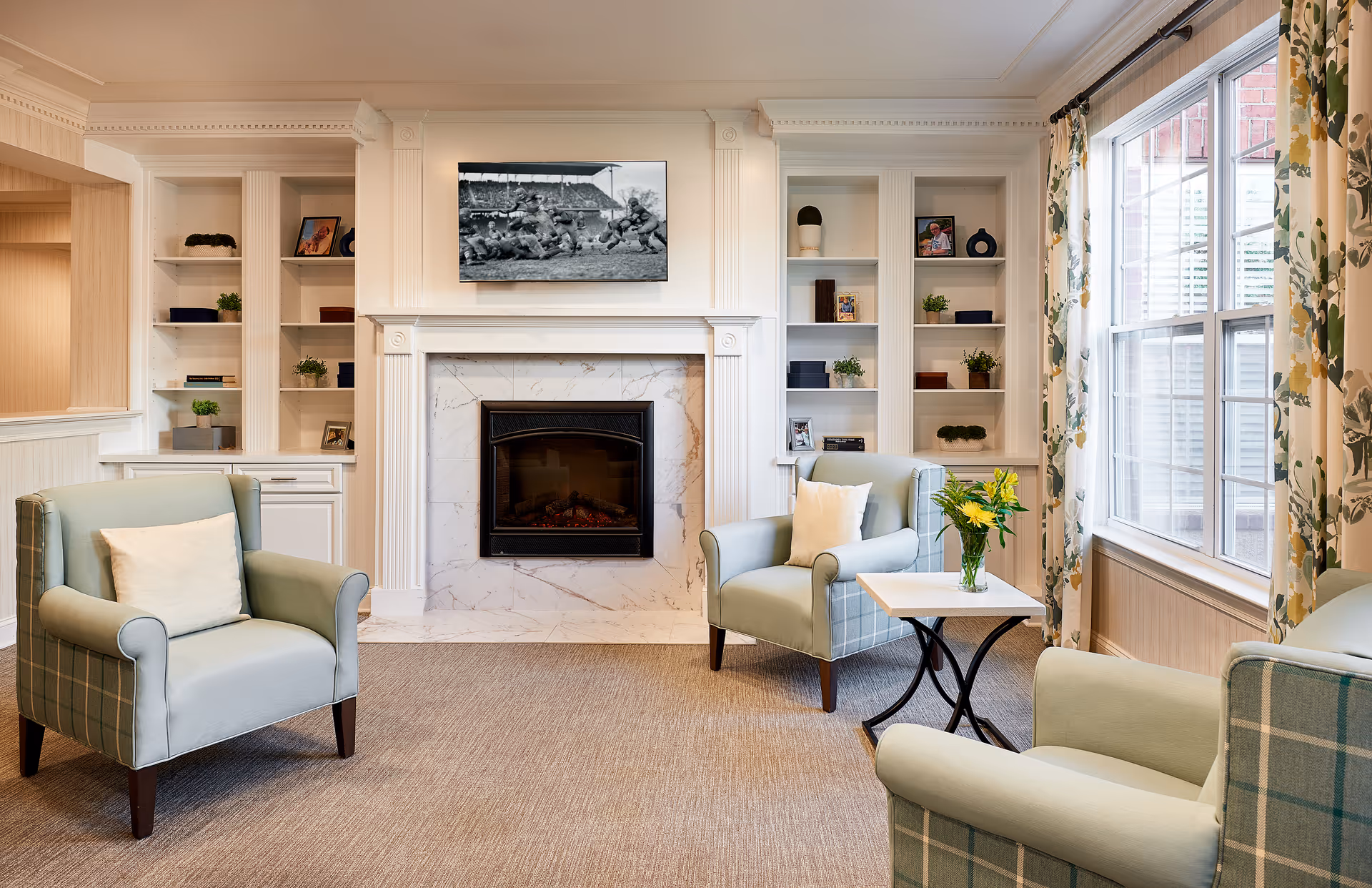 A cozy living room with three light blue armchairs arranged around a small white table with a vase of yellow flowers. The room features a white marble fireplace with a black and white photo above it, built-in white shelves with decorative items, and large windows with floral curtains letting in natural light.