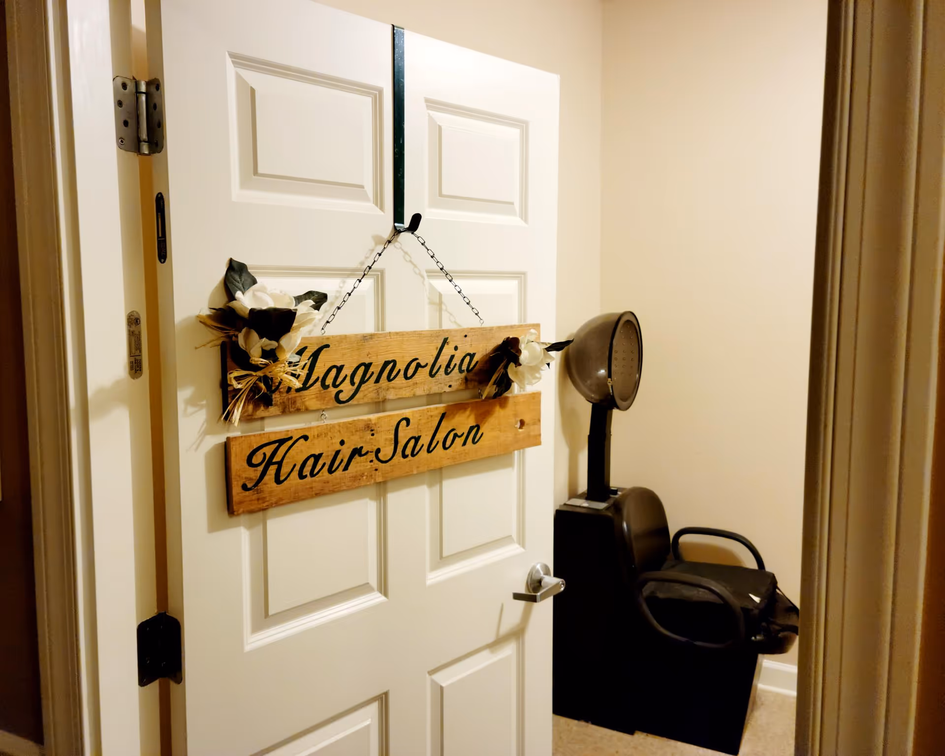 Open white door with a wooden 'Magnolia Hair Salon' sign hanging, revealing a salon chair and hooded hair dryer inside a small room.