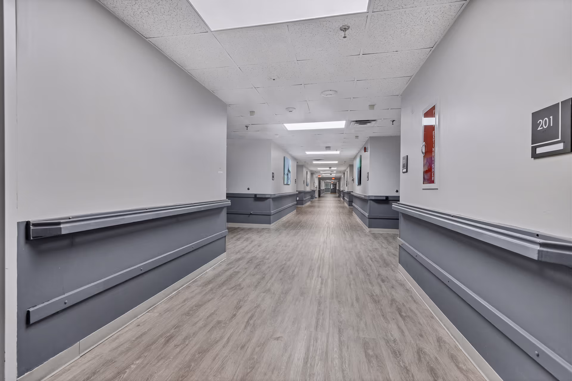 A long, empty hallway in a senior living facility with light gray walls, dark gray wainscoting, and wood-patterned flooring. The ceiling has recessed lighting and ceiling tiles. Room number 201 is visible on the right wall.