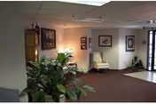 Lobby-style interior with potted plants, framed artwork, a yellow armchair, and an entry door under a drop ceiling.