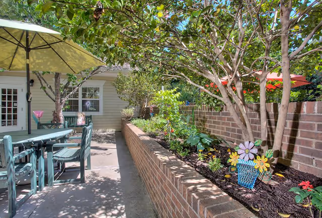 Outdoor patio area with a green wooden table and chairs under a large yellow umbrella. There is a brick planter filled with various plants and flowers along the right side, and a brick wall behind it. Trees provide shade over the area, and a building with windows and a door is visible in the background.