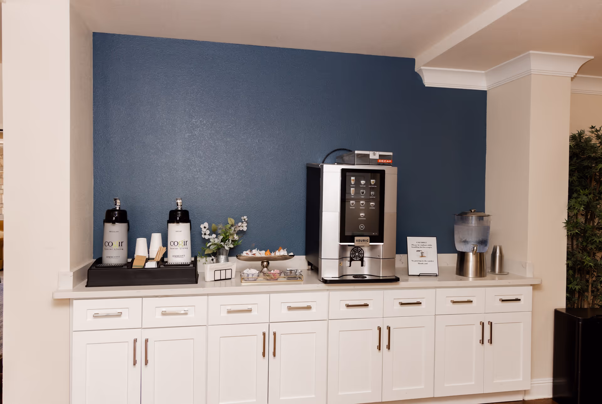 A beverage station with white cabinets and a blue accent wall. The station includes two large coffee dispensers labeled Cogir Senior Living, a Keurig coffee machine, a water dispenser, cups, stirrers, and small decorative plants and snacks.
