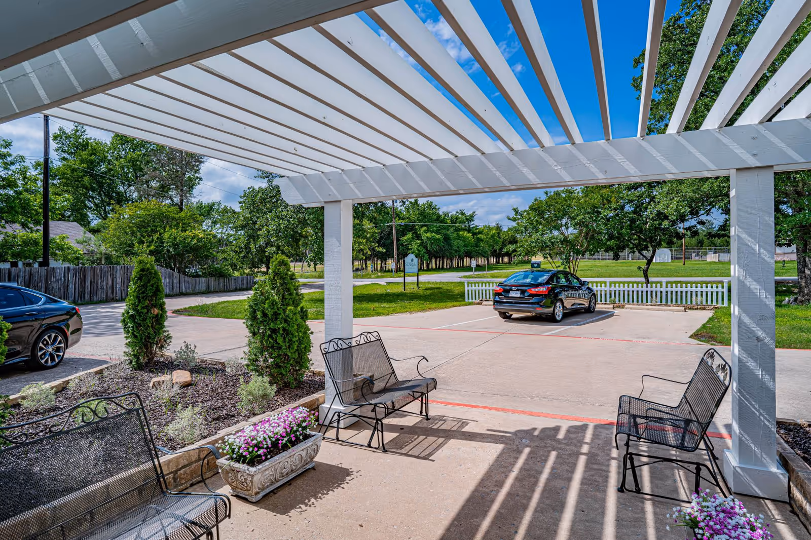 Outdoor seating area with metal benches and flower planters under a white pergola. In the background, there is a parking lot with two cars and green trees under a blue sky with some clouds.