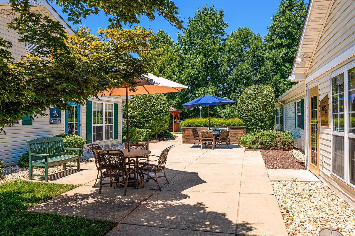 Sunny outdoor courtyard at a memory-care community with patio tables, umbrellas, benches and landscaped greenery between buildings.