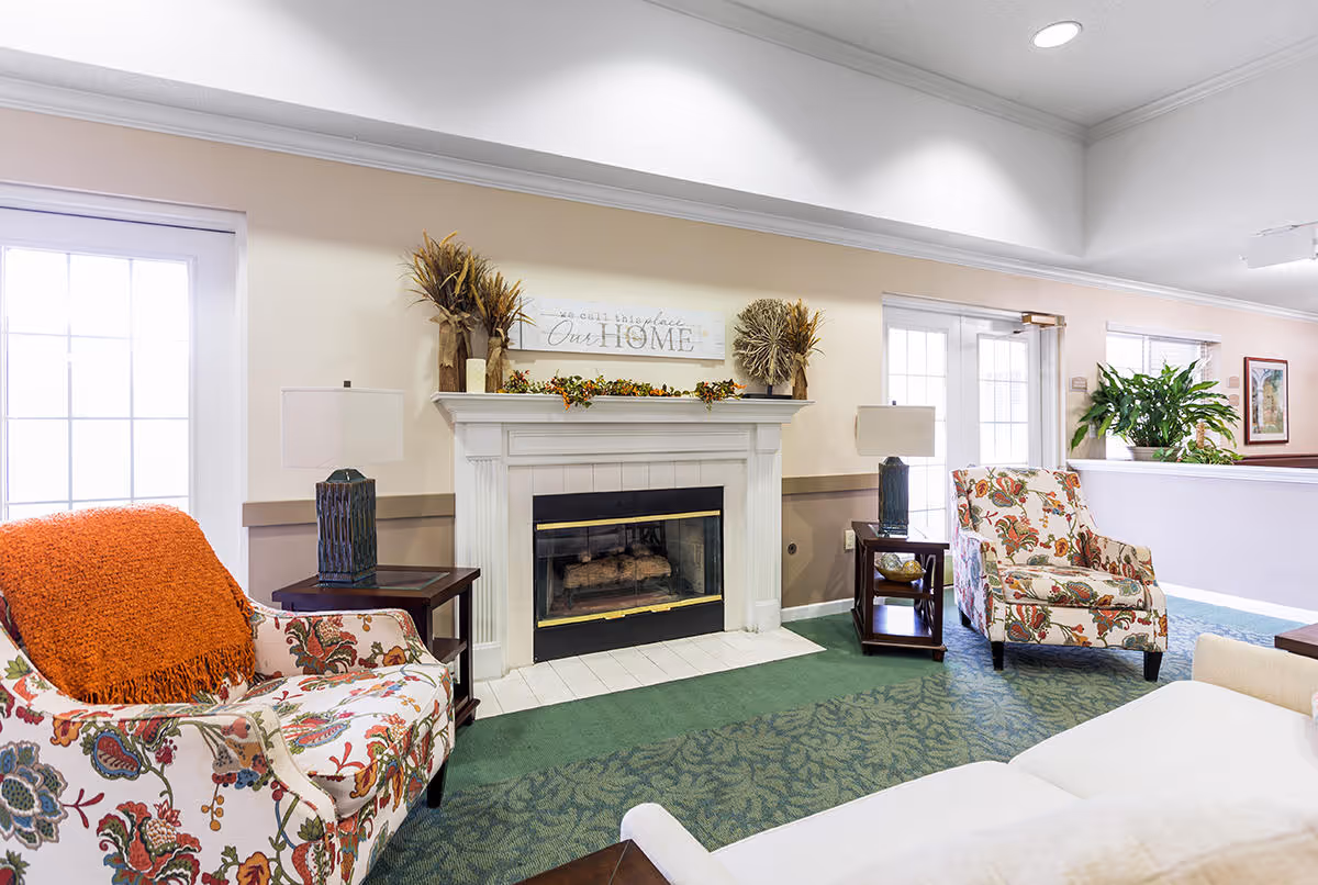 A cozy living room area in a senior living facility featuring a white fireplace with decorative plants and a sign above it that reads 'We call this place Our Home'. The room has two floral patterned armchairs, one with an orange throw blanket, two side tables with lamps, a white couch, green carpet, and large windows letting in natural light.