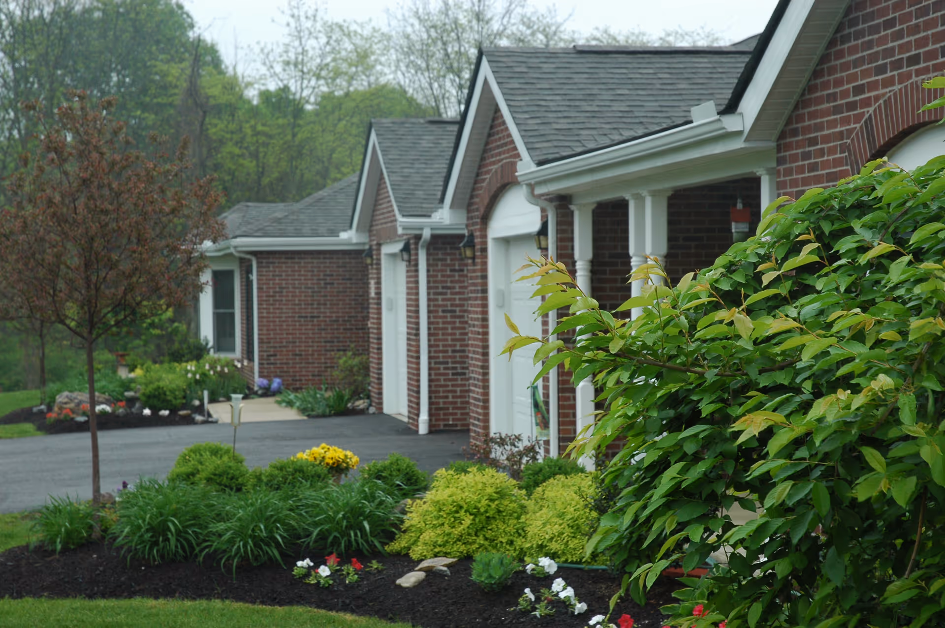 View of a row of brick residential buildings with white garage doors and columns, surrounded by well-maintained landscaping including green shrubs, flowering plants, and a small tree, with a paved driveway in front.