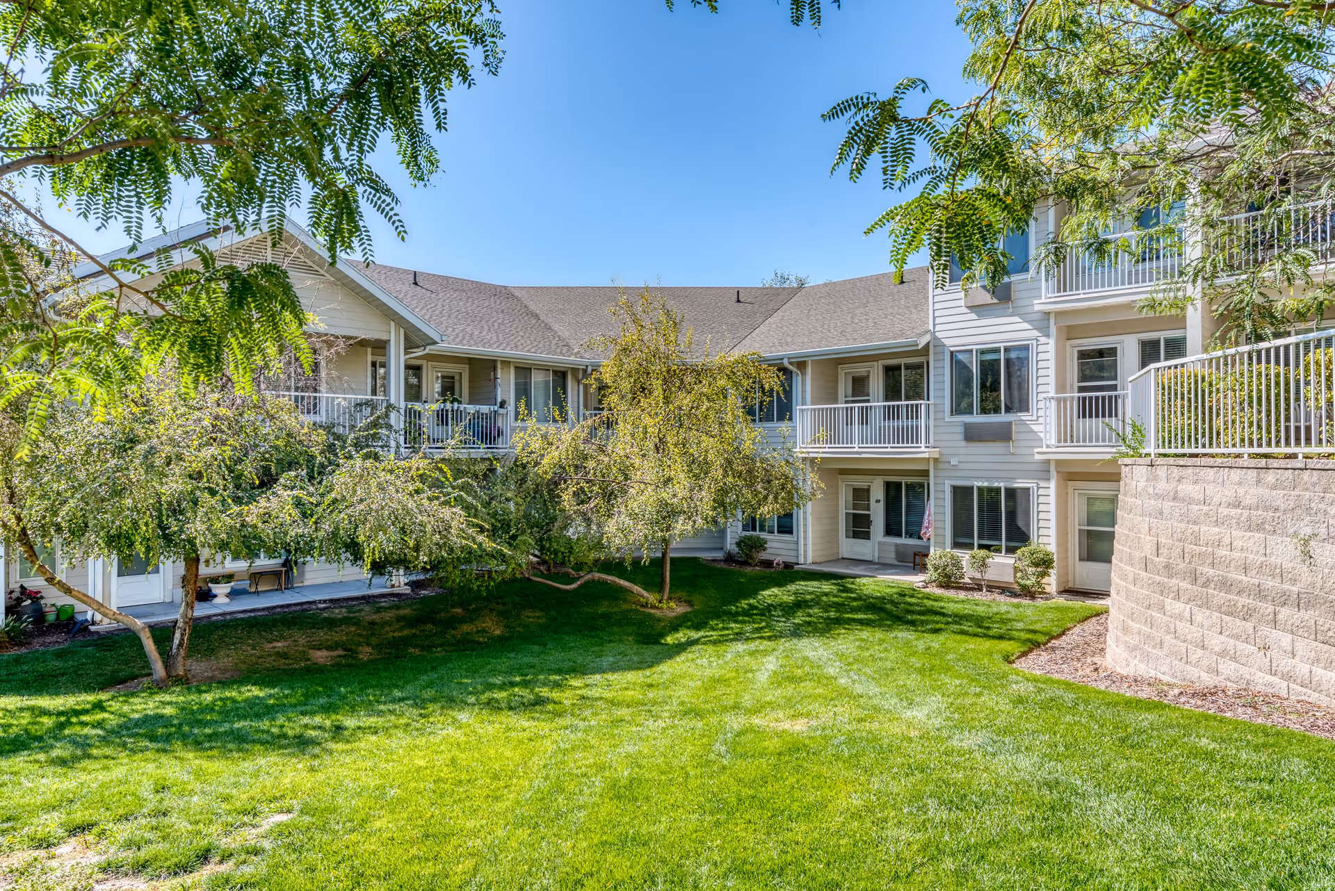 Exterior view of a senior living facility building with multiple balconies and windows, surrounded by green grass, trees, and landscaping under a clear blue sky.