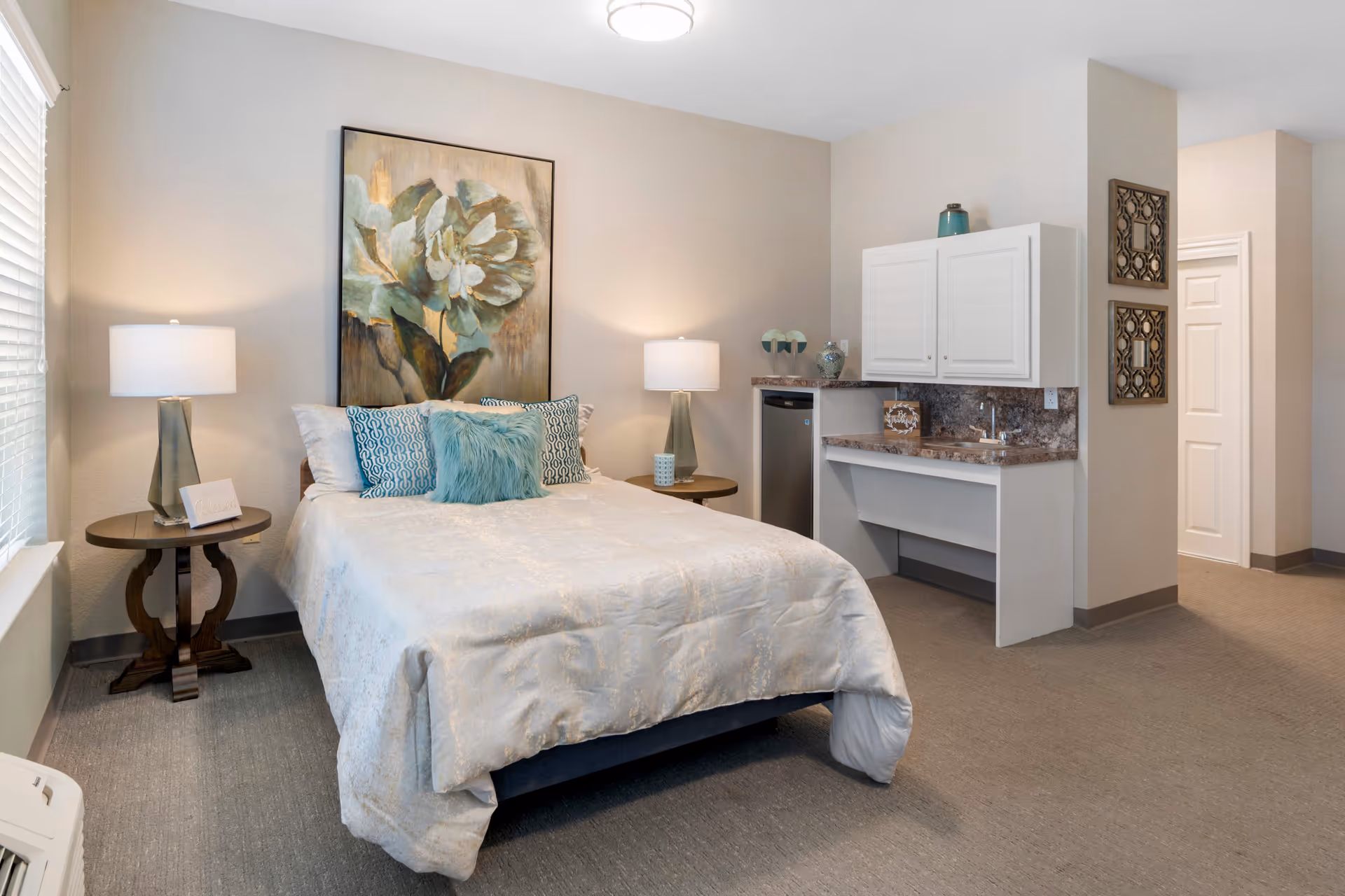 A furnished bedroom in a senior living unit with a bed, bedside tables and lamps, floral artwork, and a small kitchenette area.