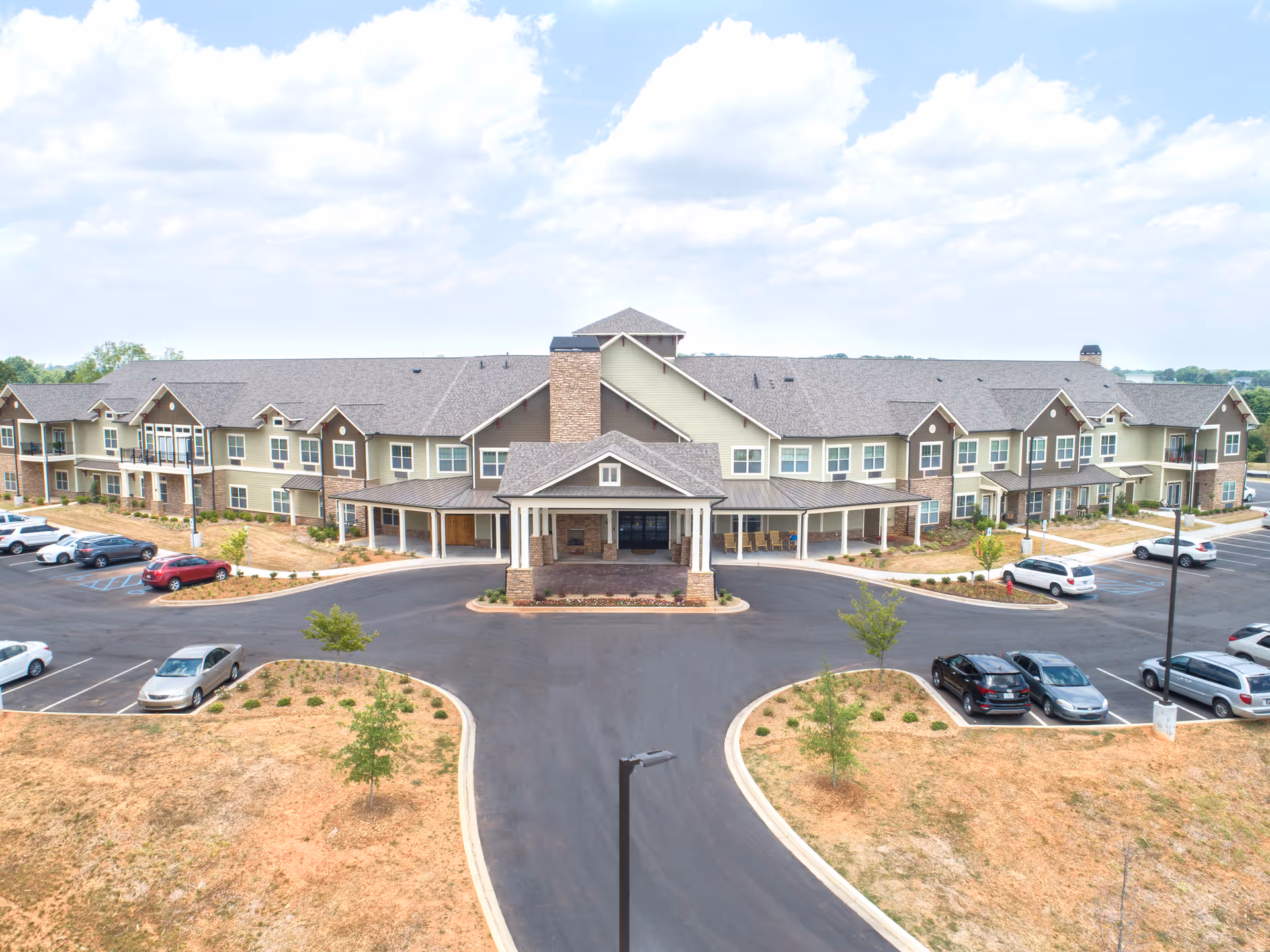 Front exterior view of Dominion Senior Living of Anderson, a large two-story building with a covered entrance, multiple windows, and a parking lot with several cars parked. The surrounding area has some small trees and landscaping with a partly cloudy sky above.