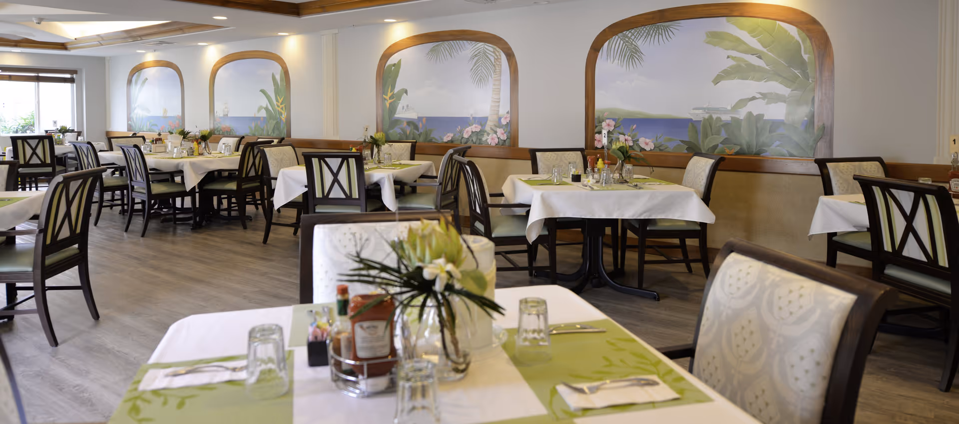 A bright and spacious dining room with multiple tables covered in white tablecloths and green placemats. Each table is set with glasses, silverware, and small floral centerpieces. The walls feature large decorative panels with tropical-themed murals, and the floor is a light wood finish. Chairs with dark wooden frames and cushioned seats surround the tables.