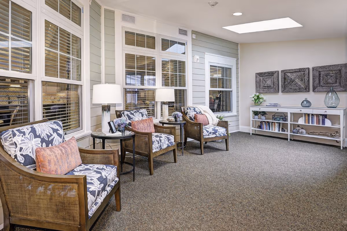 A cozy sitting area with three wooden armchairs featuring floral cushions and orange accent pillows, two side tables with lamps and small flower arrangements, large windows with white blinds, a white console table with books and decorative items, and three wooden wall art pieces above the console.