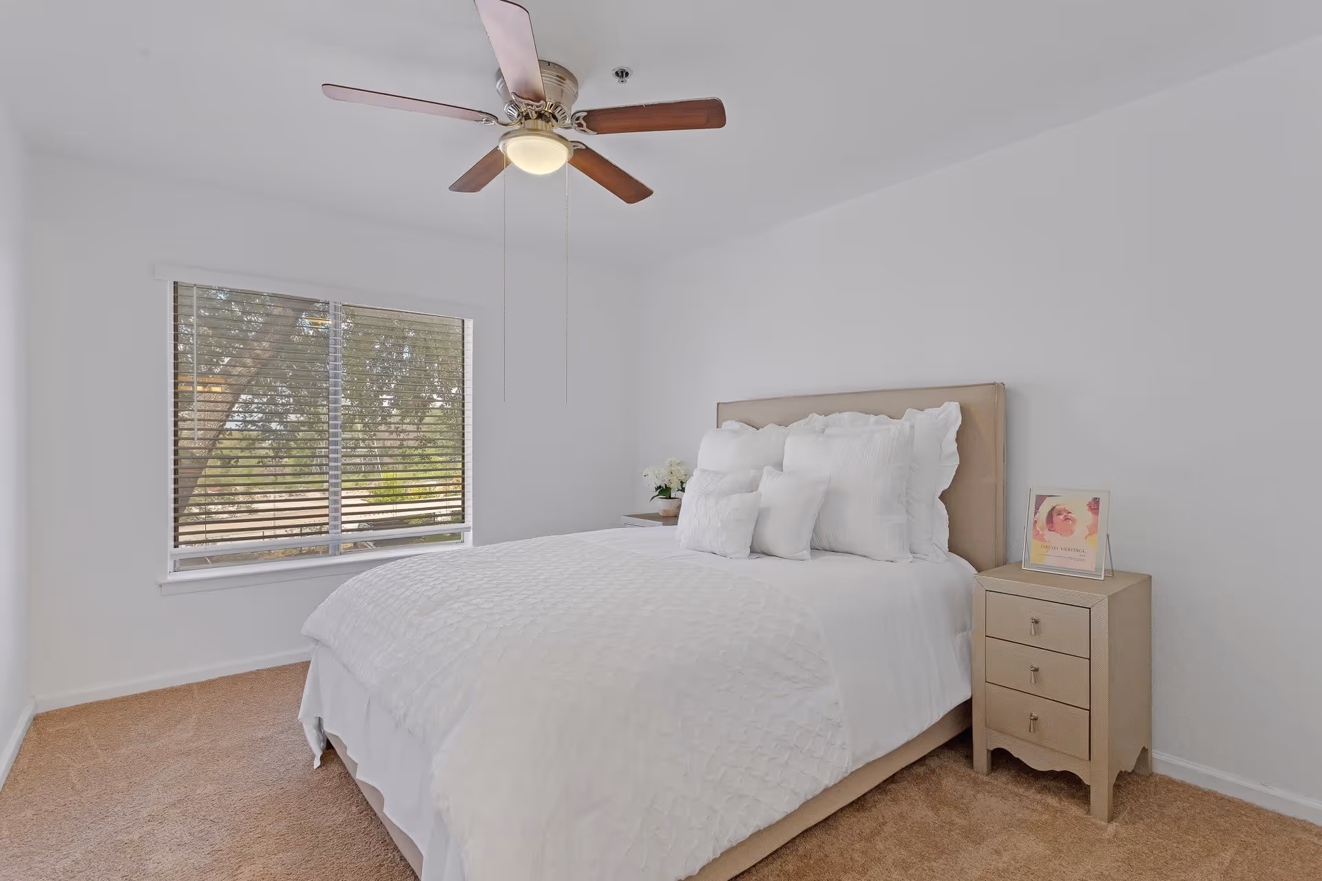 A bright bedroom with a large window covered by blinds, a ceiling fan with wooden blades, a bed with white bedding and multiple pillows, and a beige nightstand with three drawers and a framed picture on top.