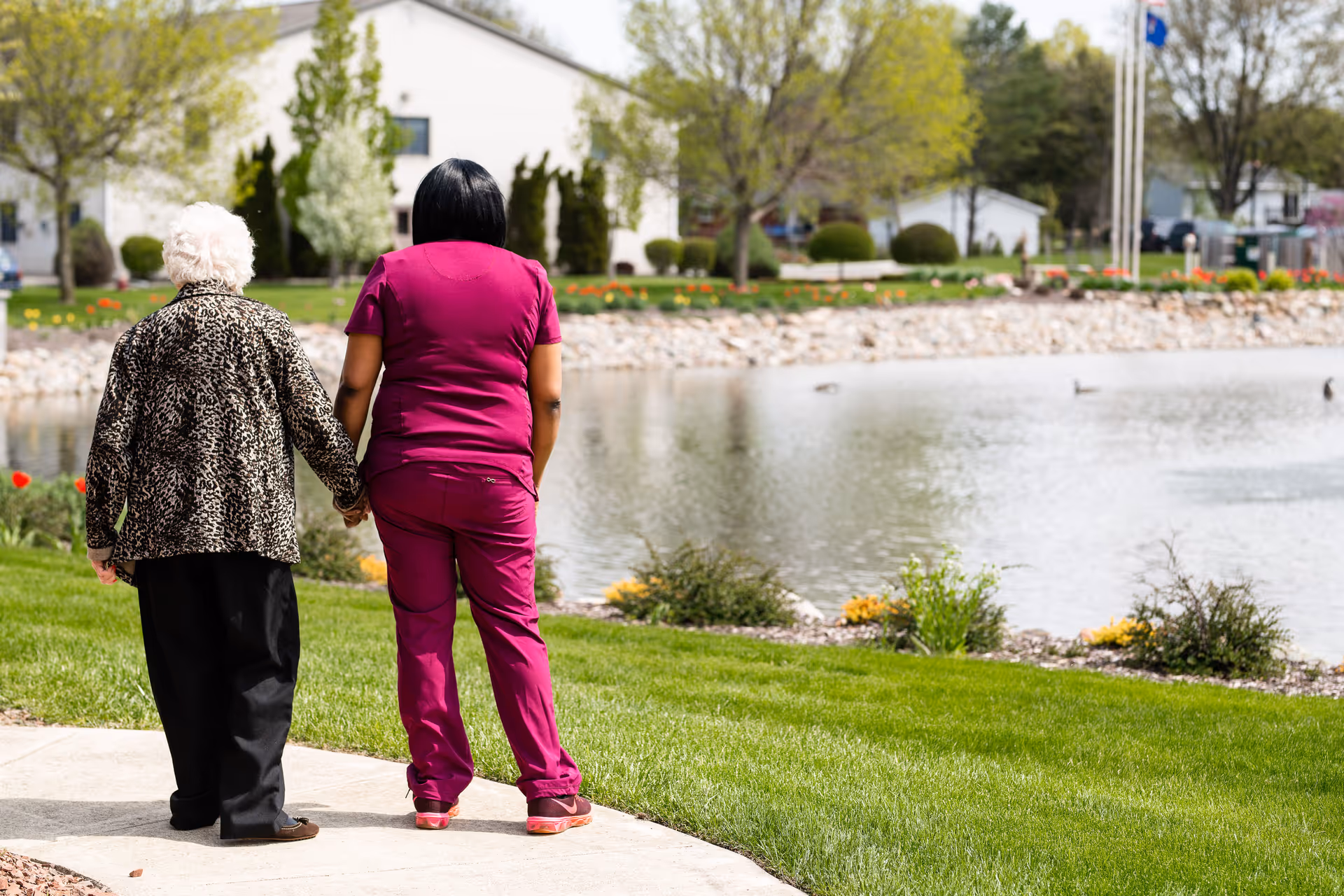 An elderly woman holding hands with a caregiver dressed in magenta scrubs as they walk along a paved path beside a pond with ducks. Trees, bushes, and a white building are visible in the background under a clear sky.
