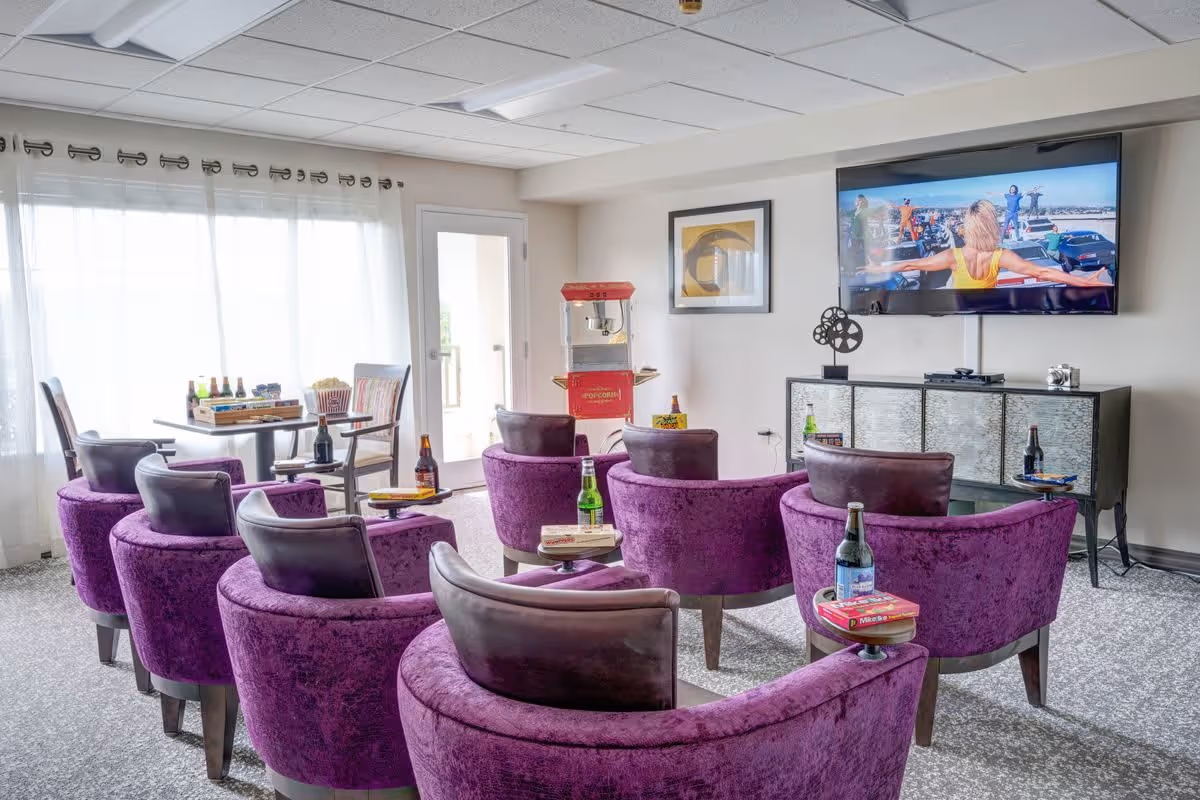 A cozy entertainment room with eight purple armchairs arranged in two rows facing a wall-mounted flat-screen TV. Each chair has a small round table attached, holding bottles and board games. There is a popcorn machine and a small table with chairs near a large window with sheer curtains, letting in natural light.