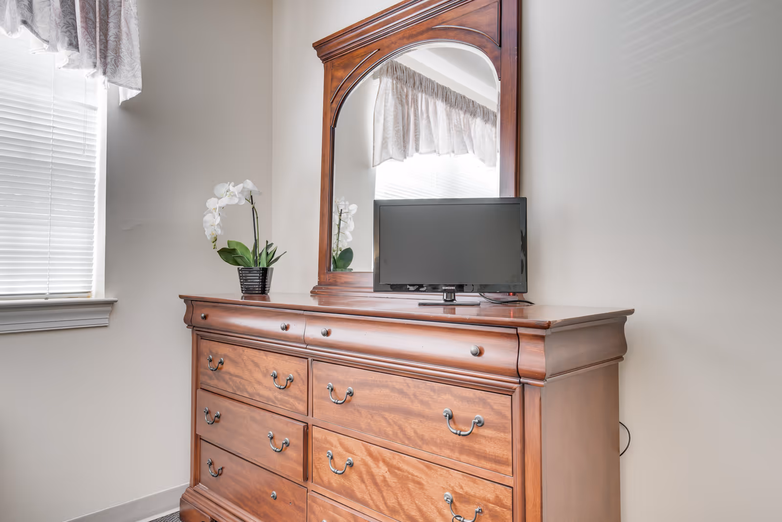 A wooden dresser with multiple drawers and a large attached mirror. On top of the dresser is a small flat-screen TV and a potted white orchid plant. A window with blinds and a valance is visible to the left.