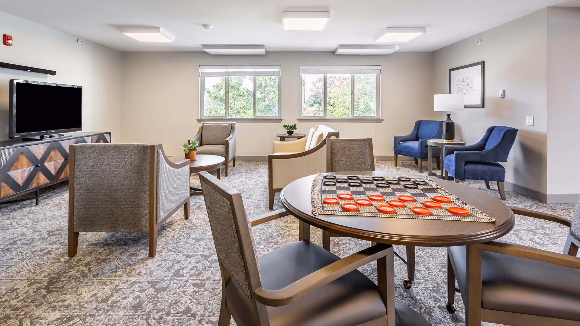 A bright and spacious living room area with multiple armchairs arranged around small tables. A round table in the foreground has a checkers game set up with red and black pieces. Large windows let in natural light, and a flat-screen TV is mounted on a patterned wooden console against one wall. The room has a neutral color palette with blue accent chairs and modern lighting fixtures on the ceiling.