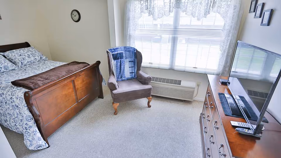 A bright bedroom with a wooden bed frame, floral bedding, a brown blanket, a cushioned armchair with a blue patchwork throw, a wooden dresser with a flat-screen TV and remote controls, a large window with lace curtains, and a wall clock.