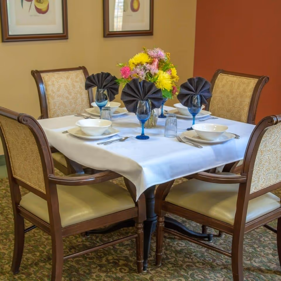A dining table set for four with a white tablecloth, blue-stemmed glasses, folded dark napkins, and a vase of colorful flowers.